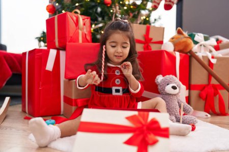 Foto de Adorable chica hispana desempacando regalo sentado junto al árbol de Navidad en casa - Imagen libre de derechos