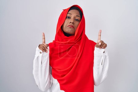 Téléchargez les photos : Jeune femme arabe portant l'écharpe traditionnelle islamique hijab pointant vers le haut regardant triste et contrarié, indiquant la direction avec les doigts, malheureux et déprimé. - en image libre de droit