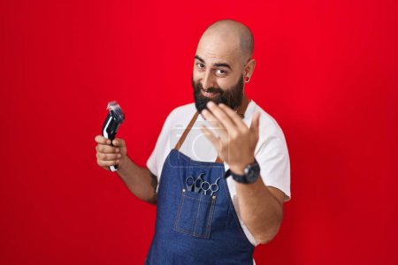 Photo for Young hispanic man with beard and tattoos wearing barber apron holding razor beckoning come here gesture with hand inviting welcoming happy and smiling - Royalty Free Image