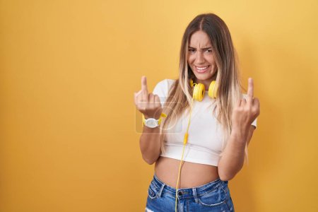 Foto de Young blonde woman standing over yellow background wearing headphones showing middle finger doing fuck you bad expression, provocation and rude attitude. screaming excited - Imagen libre de derechos
