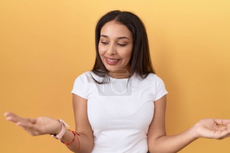Photo for Young arab woman wearing casual white t shirt over yellow background smiling showing both hands open palms, presenting and advertising comparison and balance - Royalty Free Image