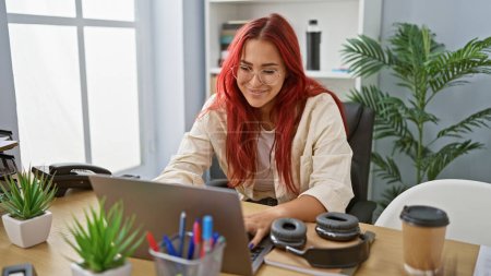 Téléchargez les photos : Femme d'affaires rousse confiante, une jeune travailleuse joyeuse, souriante alors qu'elle travaille en ligne à l'aide de son ordinateur portable dans l'élégant intérieur du bureau - en image libre de droit