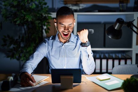 Téléchargez les photos : Young hispanic man working at the office at night angry and mad raising fist frustrated and furious while shouting with anger. rage and aggressive concept. - en image libre de droit