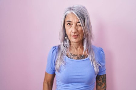 Téléchargez les photos : Femme d'âge moyen avec des tatouages debout sur fond rose clin d'oeil en regardant l'appareil photo avec une expression sexy, gai et heureux visage. - en image libre de droit