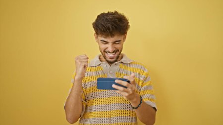 Téléchargez les photos : Jeune homme arabe confiant célébrant joyeusement sa victoire de jeu, souriant tout en jouant à un jeu vidéo sur son smartphone sur fond jaune isolé - en image libre de droit