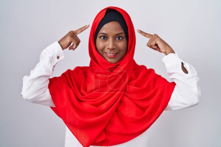 Téléchargez les photos : Jeune femme arabe portant l'écharpe traditionnelle islamique hijab souriant pointant vers la tête avec les deux doigts de la main, grande idée ou pensée, bonne mémoire - en image libre de droit