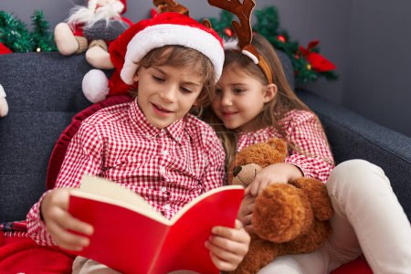 Téléchargez les photos : Frère et sœur lisant le livre assis sur le canapé par la décoration de Noël à la maison - en image libre de droit