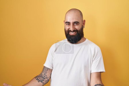 Foto de Joven hombre hispano con barba y tatuajes de pie sobre fondo amarillo sonriente alegre con los brazos abiertos como bienvenida amistosa, saludos positivos y confiados - Imagen libre de derechos
