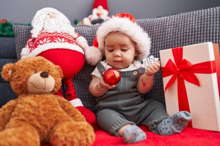 Photo for Adorable hispanic toddler sitting on sofa wearing christmas hat at home - Royalty Free Image