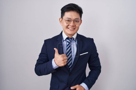 Téléchargez les photos : Jeune homme asiatique portant un costume d'affaires et une cravate faisant des pouces heureux geste avec la main. approuvant l'expression regardant la caméra montrant le succès. - en image libre de droit