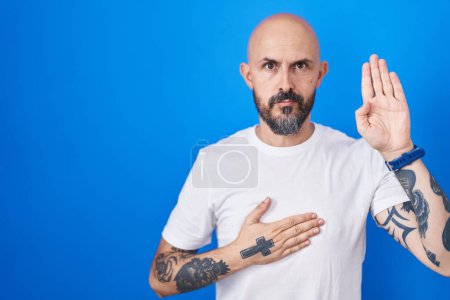 Foto de Hombre hispano con tatuajes de pie sobre fondo azul jurando con la mano en el pecho y la palma abierta, haciendo un juramento de lealtad promesa - Imagen libre de derechos