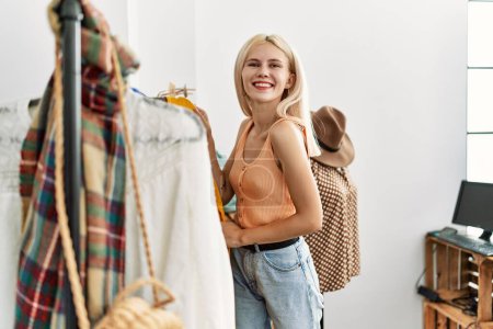 Photo for Young blonde woman customer choosing clothes smiling at clothing store - Royalty Free Image