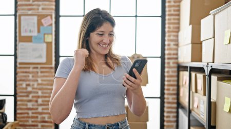 Téléchargez les photos : Jeune femme blonde ecommerce travailleur d'affaires en utilisant un smartphone avec expression gagnante au bureau - en image libre de droit