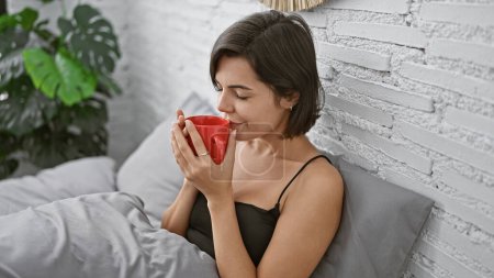Foto de Mujer hispana joven relajada sentada cómodamente en su cama, oliendo el aroma encantador del café de la mañana. hermosa mujer en su dormitorio disfrutando de su bebida con una sonrisa segura. - Imagen libre de derechos