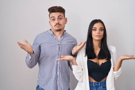 Photo for Young hispanic couple standing over white background clueless and confused expression with arms and hands raised. doubt concept. - Royalty Free Image