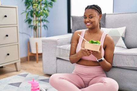 Photo for African american woman with braids eating salad after working out at home looking positive and happy standing and smiling with a confident smile showing teeth - Royalty Free Image