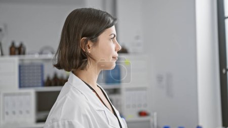 Photo for Focused young and beautiful hispanic female scientist with a serious face, looking off to the side in her busily working lab, surrounded by the hustle of science and medicine. - Royalty Free Image