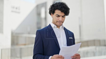 Téléchargez les photos : Jeune homme d'affaires latino-américain debout avec expression détendue document de lecture à la rue - en image libre de droit