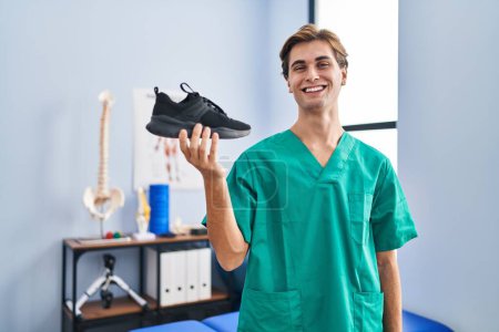Photo for Young man working at physiotherapy clinic holding shoe looking positive and happy standing and smiling with a confident smile showing teeth - Royalty Free Image