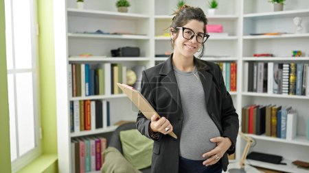 Photo for Young pregnant woman teacher holding clipboard touching belly at library university - Royalty Free Image