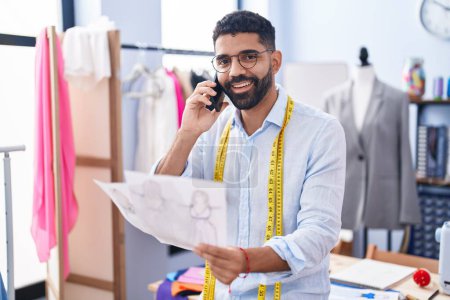 Téléchargez les photos : Young arab man tailor talking on smartphone looking clothing design at tailor shop - en image libre de droit