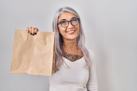 Foto de Mujer de mediana edad con pelo gris sosteniendo llevar bolsa de papel mirando positiva y feliz de pie y sonriendo con una sonrisa confiada mostrando los dientes - Imagen libre de derechos