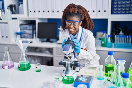 Foto de Científica afroamericana sonriendo confiada usando microscopio en laboratorio - Imagen libre de derechos