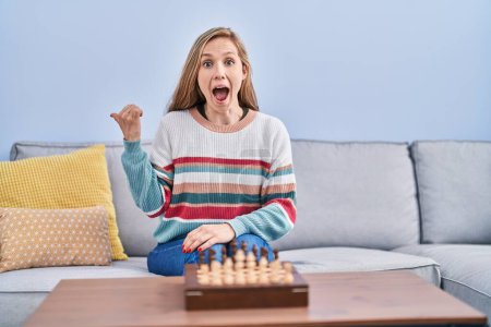 Photo for Young blonde woman playing chess sitting on the sofa pointing thumb up to the side smiling happy with open mouth - Royalty Free Image
