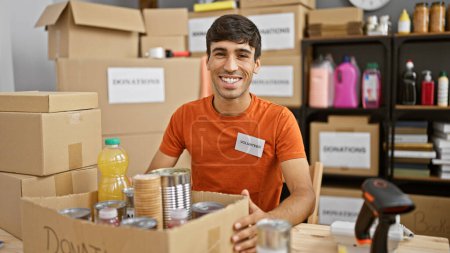 Photo for Heartwarming portrait of a young, handsome hispanic man, smiling confidently while volunteering at a charity center, sitting at a table, filled with donations in a vibrant community room. - Royalty Free Image