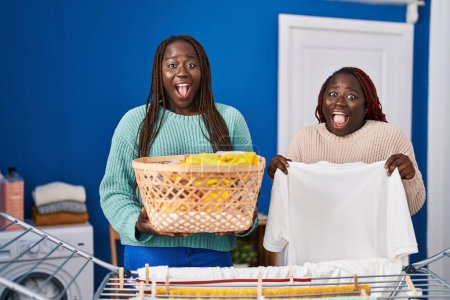 Téléchargez les photos : Deux femmes africaines accrochant des vêtements à la corde à linge célébrant fou et étonné pour le succès avec les yeux ouverts hurlant excité. - en image libre de droit