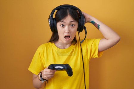 Téléchargez les photos : Chinois jeune femme jouer jeu vidéo tenant contrôleur fou et effrayé avec les mains sur la tête, peur et surpris de choc avec la bouche ouverte - en image libre de droit