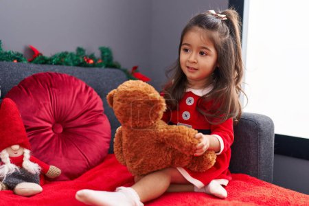 Foto de Adorable chica hispana jugando con oso de peluche sentado en el sofá por la decoración de Navidad en casa - Imagen libre de derechos