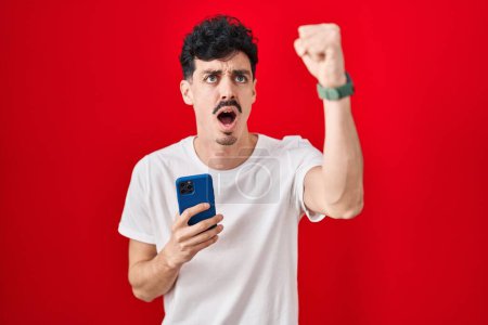 Photo for Hispanic man using smartphone over red background angry and mad raising fist frustrated and furious while shouting with anger. rage and aggressive concept. - Royalty Free Image