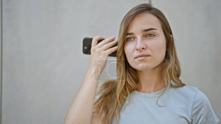 Photo for Urban loneliness, beautiful, young caucasian woman, attractively casual yet worried, standing outside on a city street, absorbing a serious voice message on her phone - Royalty Free Image