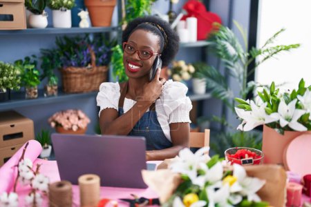 Foto de African american woman florist talking on smartphone using laptop at florist store - Imagen libre de derechos
