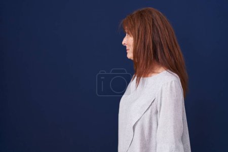 Téléchargez les photos : Femme d'âge moyen debout sur fond bleu regardant vers le côté, pose de profil relax avec visage naturel avec sourire confiant. - en image libre de droit