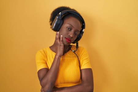 Téléchargez les photos : Femme africaine avec les cheveux bouclés debout sur fond jaune portant des écouteurs pensant fatigué et ennuyé avec des problèmes de dépression avec les bras croisés. - en image libre de droit