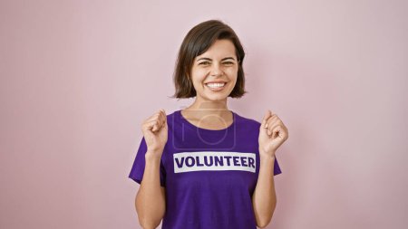 Téléchargez les photos : Jeune femme hispanique confiante célébrant sa victoire en tant que bénévole, portant un t-shirt uniforme et une coiffure courte, riant de joie sur un fond rose isolé - en image libre de droit