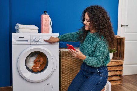 Photo for Young beautiful hispanic woman using smartphone turning on washing machine at laundry room - Royalty Free Image