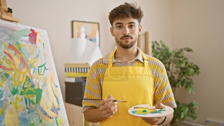 Photo for Young arab man artist holding paintbrush and palette standing at art studio - Royalty Free Image