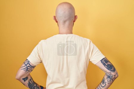 Foto de Hombre hispano con tatuajes de pie sobre fondo amarillo de pie hacia atrás mirando hacia otro lado con los brazos en el cuerpo - Imagen libre de derechos