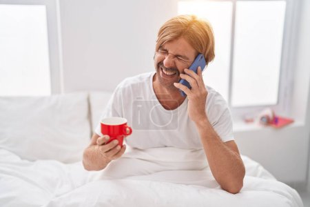 Photo for Young blond man talking on smartphone drinking coffee at bedroom - Royalty Free Image