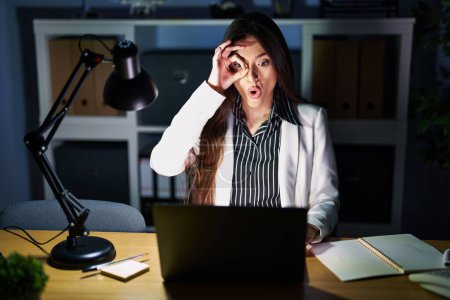 Téléchargez les photos : Jeune femme brune travaillant au bureau la nuit avec un ordinateur portable faisant un geste correct choqué par le visage surpris, les yeux regardant à travers les doigts. expression incrédule. - en image libre de droit