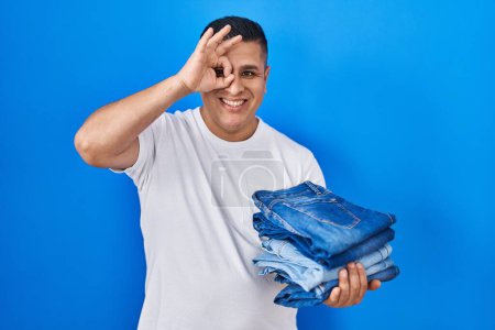 Téléchargez les photos : Jeune homme hispanique tenant pile de jeans pliés souriant heureux faisant ok signe avec la main sur l'oeil regardant à travers les doigts - en image libre de droit