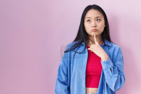 Foto de Joven asiático mujer de pie sobre rosa fondo pensamiento concentrado acerca de duda con dedo en barbilla y mirando arriba preguntando - Imagen libre de derechos