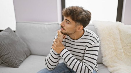 Photo for Worried young arab man with a beard, sitting on his living room sofa at home, seriously expressing doubt and sadness as he nervously ponders a problem indoors. - Royalty Free Image