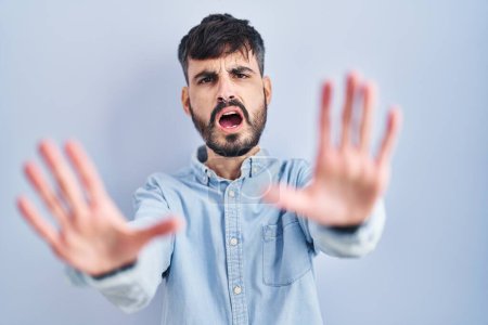 Foto de Joven hombre hispano con barba de pie sobre fondo azul haciendo gesto de stop con palmas de las manos, expresión de enojo y frustración - Imagen libre de derechos