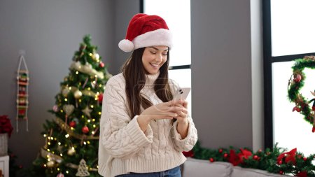 Photo for Young beautiful hispanic woman using smartphone standing by christmas tree at home - Royalty Free Image