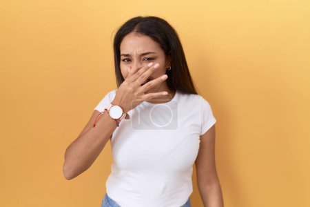 Téléchargez les photos : Jeune femme arabe portant un t-shirt blanc décontracté sur fond jaune sentant quelque chose de puant et dégoûtant, odeur intolérable, retenant la respiration avec les doigts sur le nez. mauvaise odeur - en image libre de droit