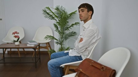 Photo for Serious-faced young hispanic man, an elegant professional, sitting pensively in a waiting room chair, casting concentrated glances around the indoor space, conveying success. - Royalty Free Image